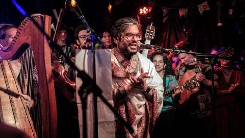 Man in a colourful outfit clapping, surrounded by people playing all sorts of instruments including a harp in the foreground